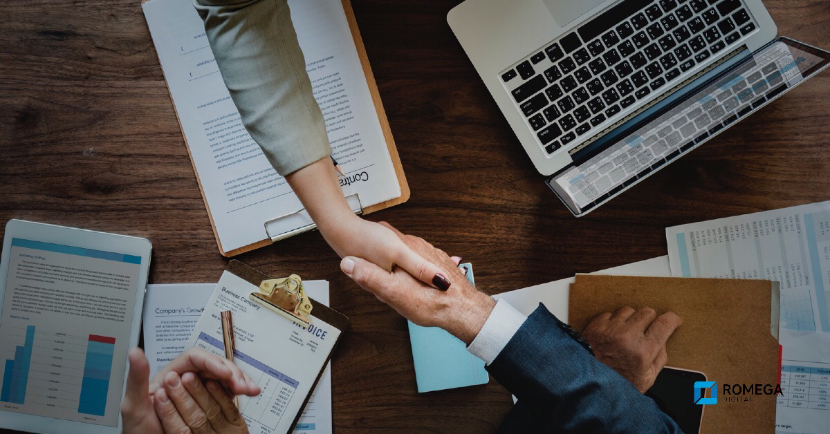 This image is of the top of a table looking down. The table is covered by different forms and official documents. Two people are reaching across the table shaking hands.